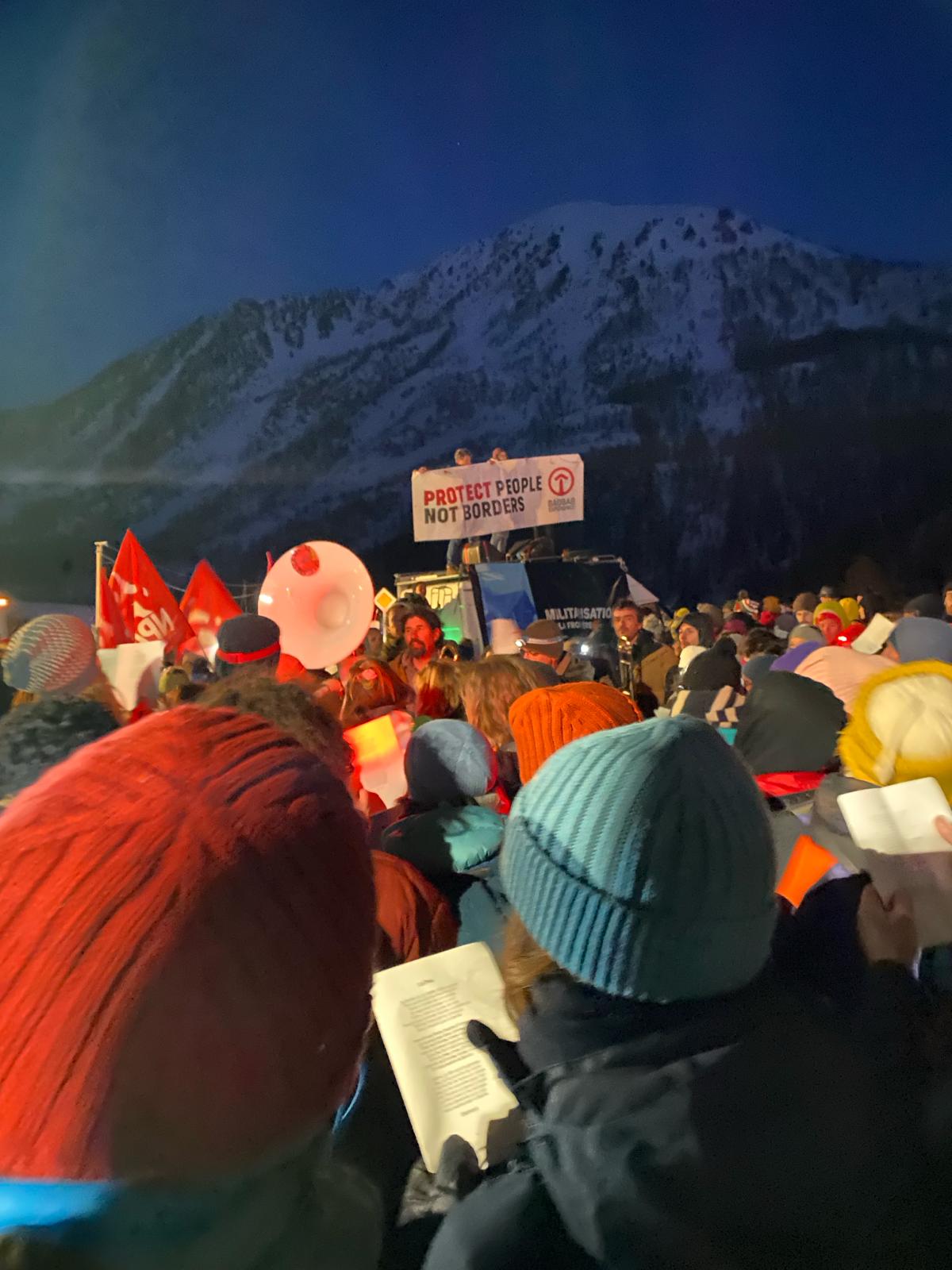 De nuit, au premier plan des tête en bonnet, qui chantent un texte militant, des drapeau flottent et une banderole "Protect People not border". Au fond de l'image une montagne enneigée