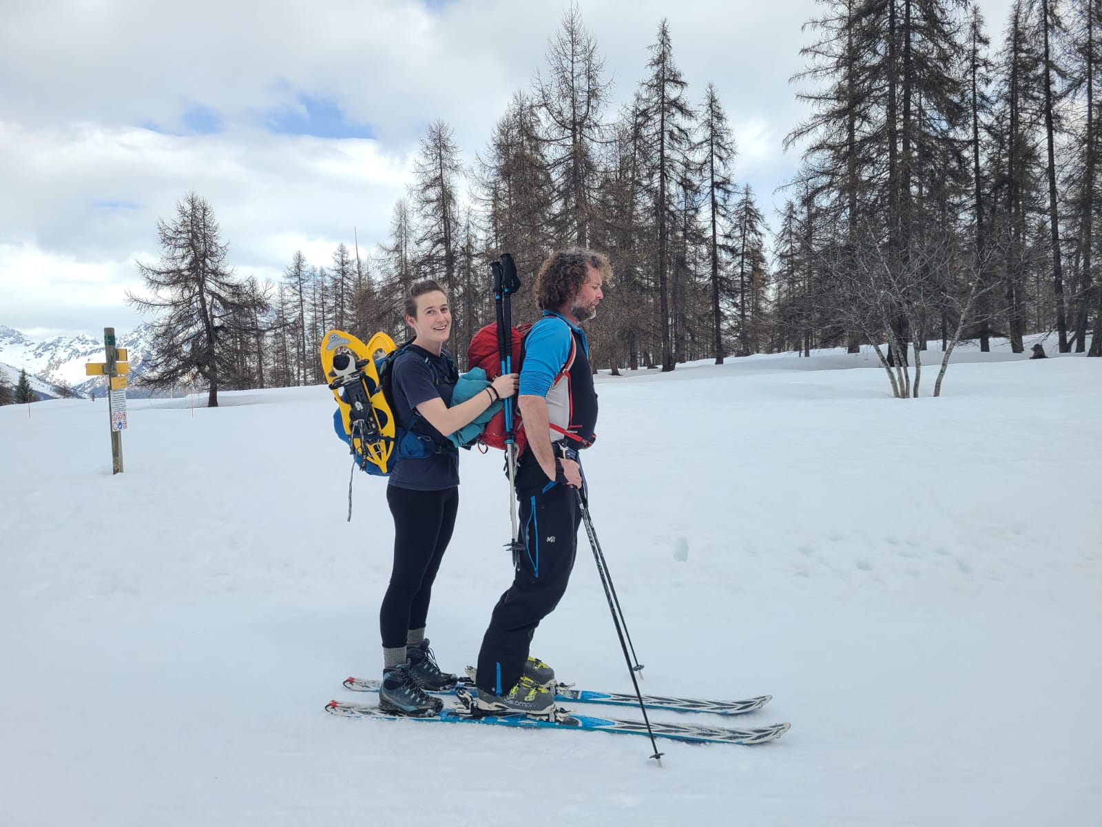 Un homme trimbale une femme sur ses propres ski, elle a placé ses raquettes sur son sac à dos, et s'accroche au sac à dos de l'homme